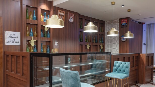 A café interior with wooden paneling, a glass display case, and two blue velvet bar stools. - Sunday Harbour Gateway Jeddah Beach Formerly Vivid Plaza Hotel (Jeddah)
