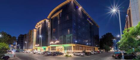 A modern hotel with a curved facade, illuminated entrance, and a parking lot with several cars.