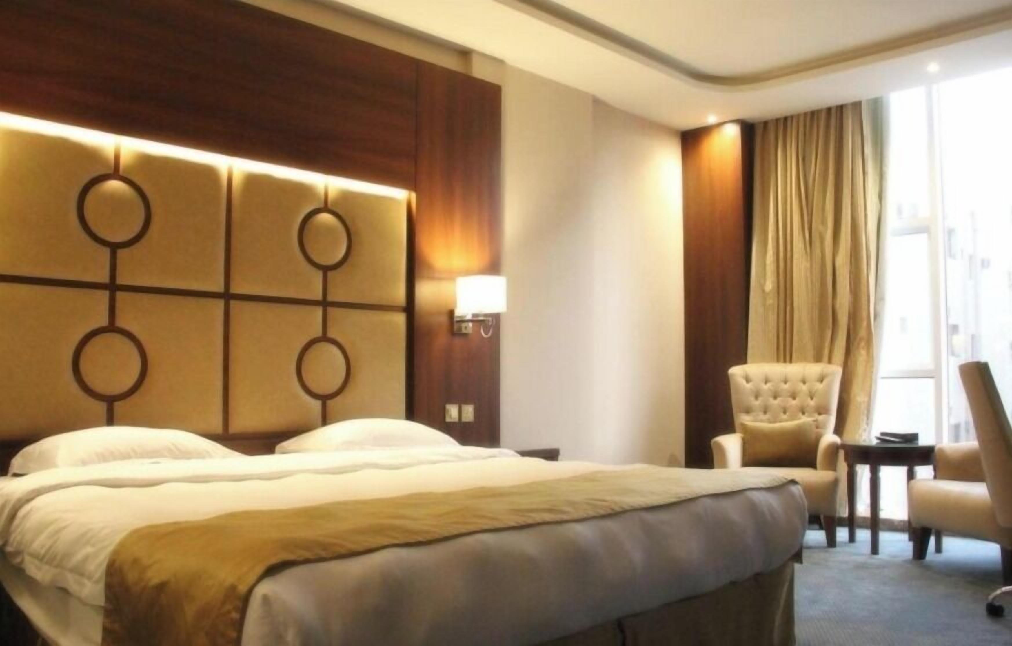 Habitación de hotel con una cama grande, cabecera con diseños circulares, mesita de noche con lámpara, una silla y vistas al exterior a través de una ventana con cortinas.