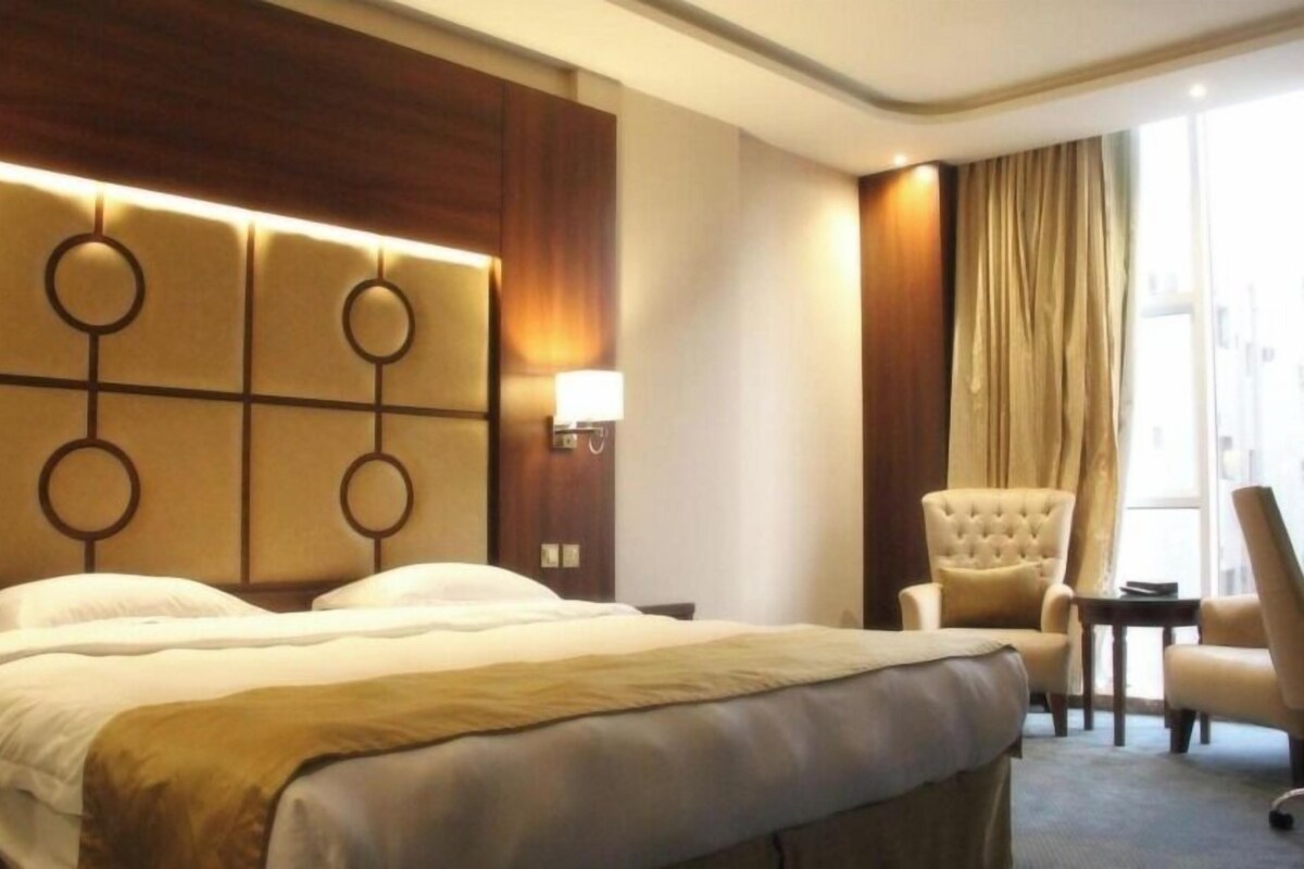 Une chambre d’hôtel avec un grand lit, une tête de lit ornée de motifs circulaires, une table de chevet avec une lampe, une chaise et une vue sur l’extérieur par une fenêtre avec des rideaux.