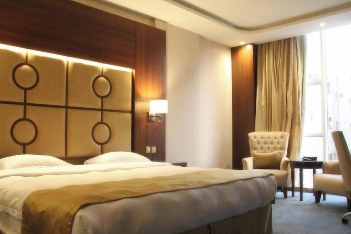 Habitación de hotel con una cama grande, cabecera con diseños circulares, mesita de noche con lámpara, una silla y vistas al exterior a través de una ventana con cortinas.