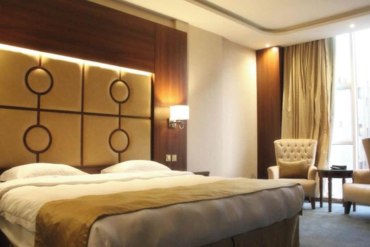 Een hotelkamer met een groot bed, een hoofdeinde met ronde patronen, een nachtkastje met een lamp, een stoel en uitzicht op buiten door een raam met gordijnen.