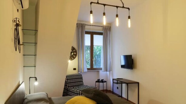 A bedroom with a bed, a chair, a TV, a window with curtains, and a ceiling light fixture. - Napoli Vesuvio By Dimorra (NAPOLI)