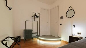 A small, well-lit room with a bed, a bedside table, a radiator, a coat rack, and a door. - Napoli Vesuvio By Dimorra (NAPOLI)