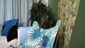 Une pièce avec un mur d’accent vert, un fauteuil orné d’un motif de feuilles tropicales, une plante en pot et un lit avec des coussins bleus et noirs.