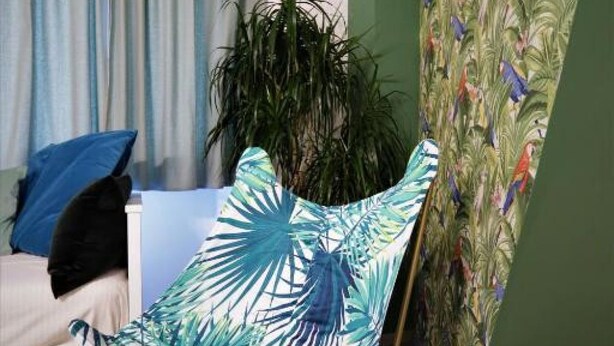 Une chambre avec un lit, un oreiller bleu, une plante et une chaise ornée d’un motif de feuilles tropicales.