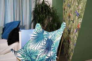 A room with a bed, a blue pillow, a plant, and a chair with a tropical leaf pattern.