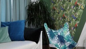 Un salon comprenant un canapé, une table ronde blanche avec deux tasses à motifs verts et blancs, un fauteuil à motifs de feuilles tropicales et un mur vert orné d’un papier peint représentant des oiseaux et des plantes.