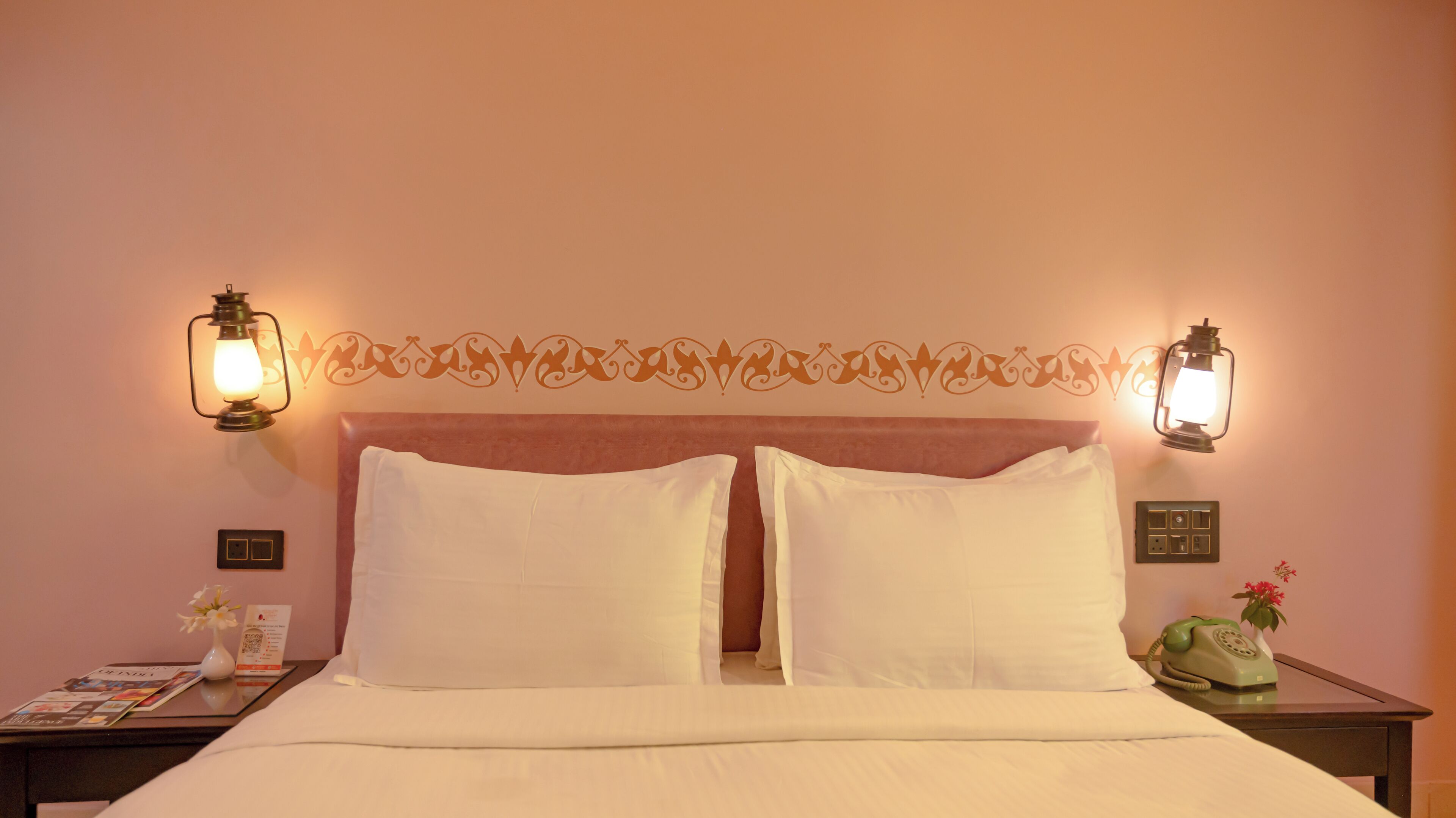Une chambre d’hôtel comprenant un lit, deux lampes fixées au mur, une tête de lit décorative, une table de chevet avec un téléphone et un petit vase avec des fleurs.