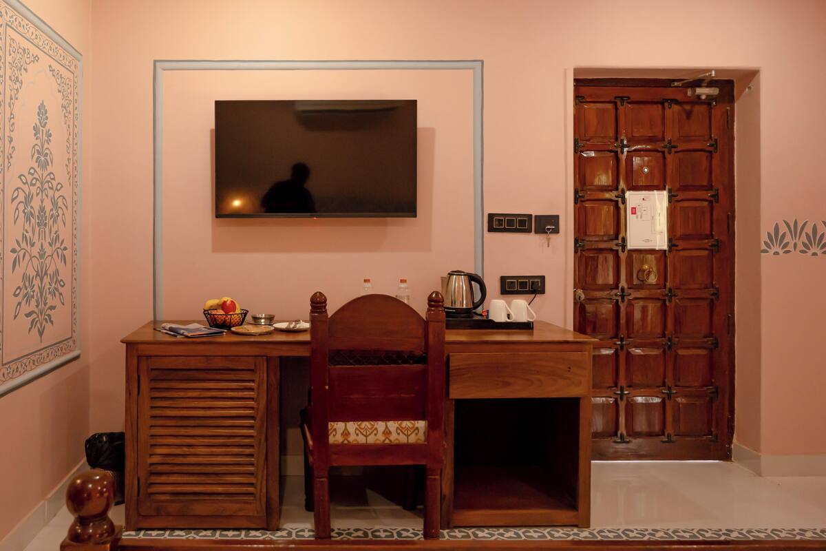A room with a wooden desk, a chair, a television mounted on the wall, a door with a lock, and a patterned wall decoration.