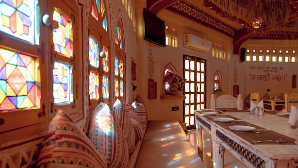 A room with a long table, patterned cushions, and stained glass windows. - NIrali Dhani Ethnic Resort (Jodhpur)