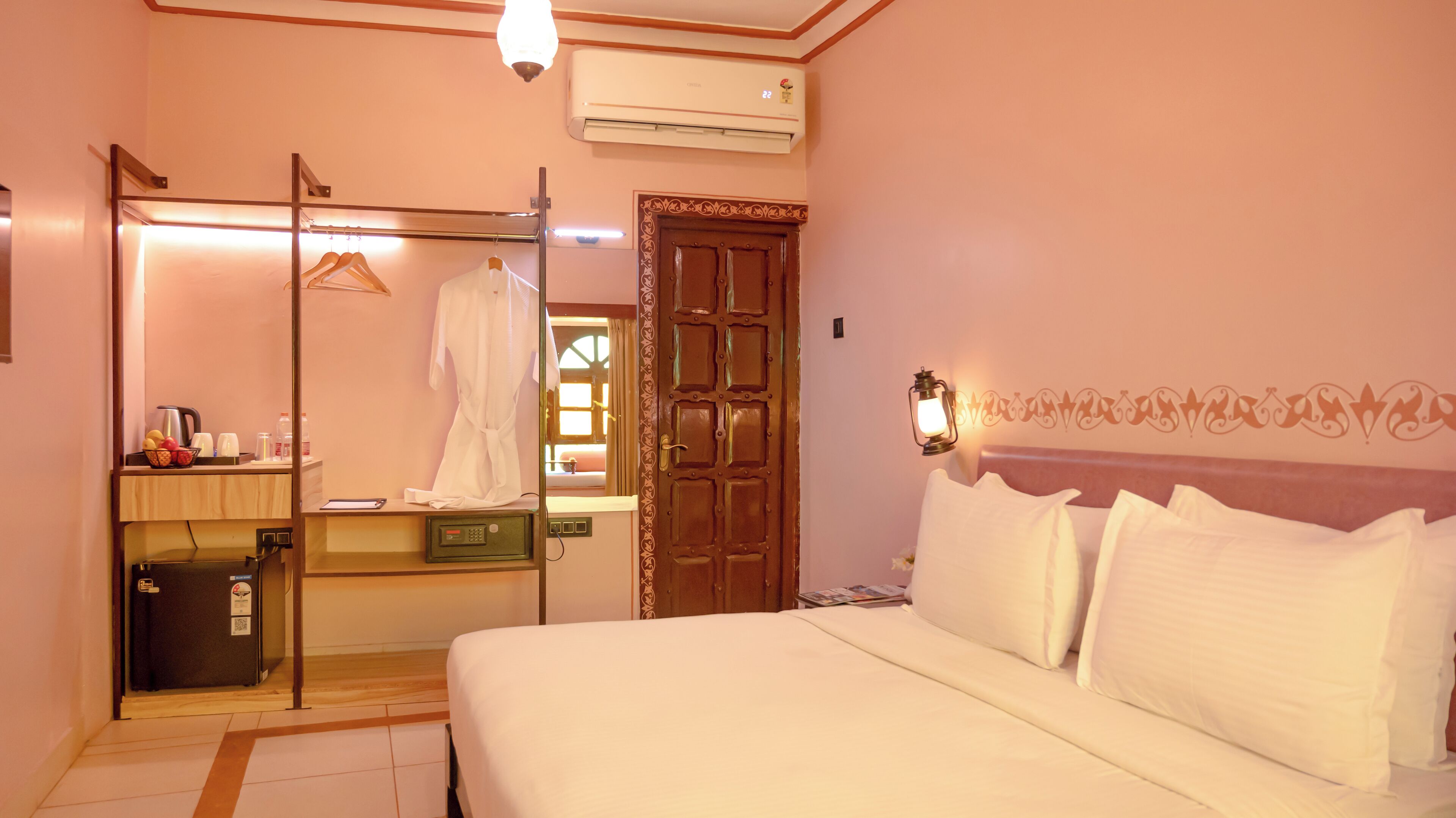 Quarto de hotel com cama, guarda-roupa, espelho, pia e pequena mesa com chaleira e fruta.