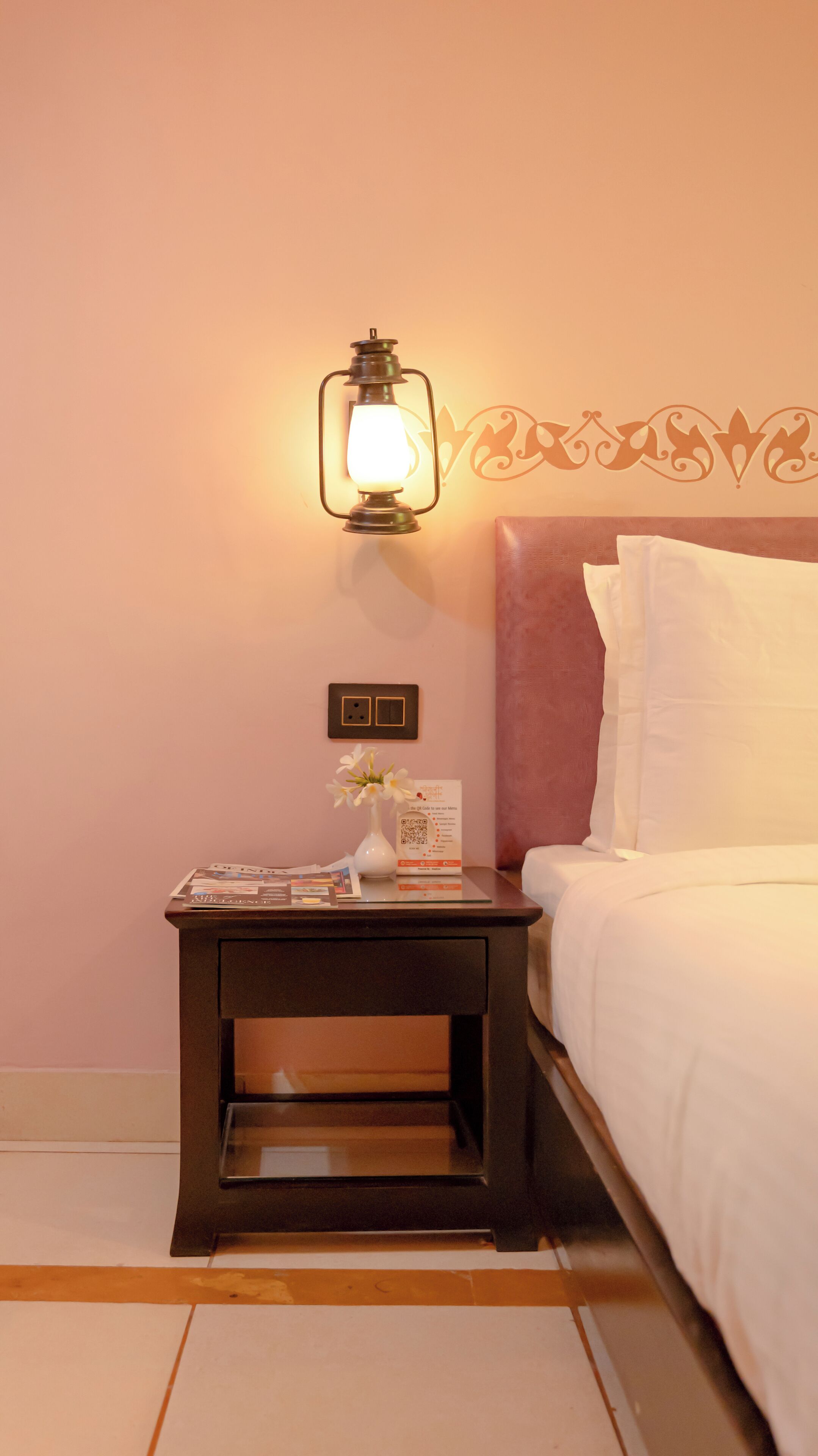 A hotel room with a bed, a nightstand with a lamp, a vase with flowers, and a wall decoration.