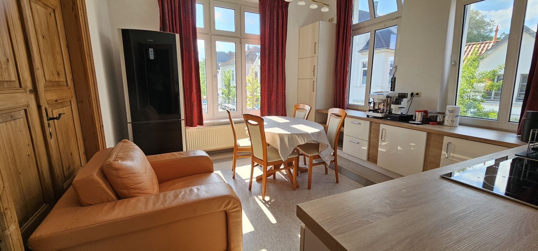 Un salon confortable avec un canapé en cuir, une table à manger avec des chaises, un coin cuisine équipé d’une cuisinière et de grandes fenêtres avec des rideaux.
