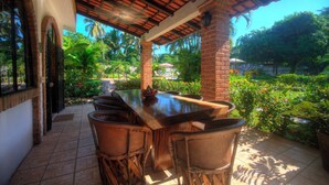 Un patio al aire libre con una mesa de madera larga y varias sillas de mimbre, rodeado de exuberante vegetación y una vista despejada del cielo.