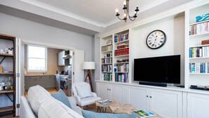 Un salon comprenant un canapé, un fauteuil, une table basse, une bibliothèque, un téléviseur et une horloge.