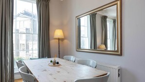 Un coin repas avec une table en bois, des chaises blanches, une lampe et un grand miroir au mur.