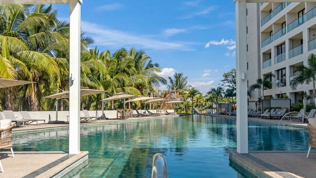 A swimming pool with lounge chairs and umbrellas, surrounded by palm trees and a multi-story building.