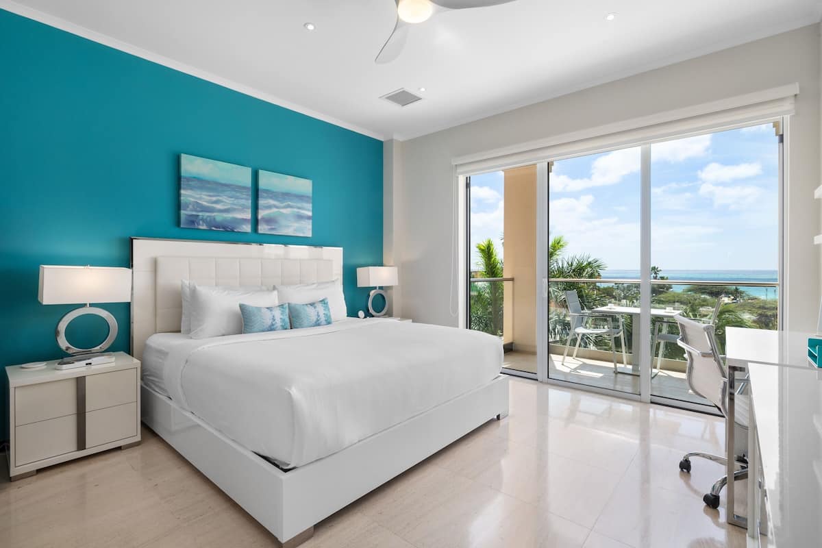 A modern bedroom with a large bed, a nightstand, a desk, and a balcony with a view of the ocean.