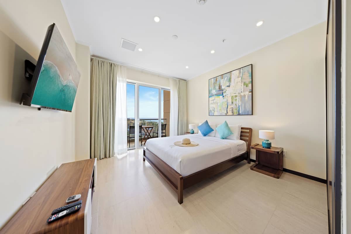 A hotel room with a bed, a TV mounted on the wall, a wooden nightstand, and a view of the outdoors through a sliding glass door.