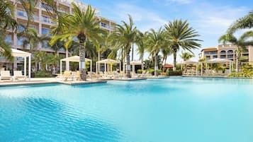 Una piscina con sillones de playa y sombrillas, rodeada de palmeras y edificios de hotel.