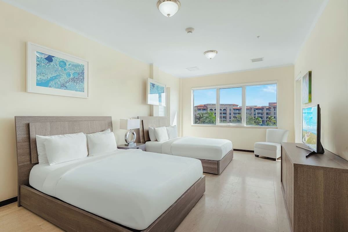 A hotel room with a large bed, a smaller bed, a desk, a chair, and a large window with a view of buildings.