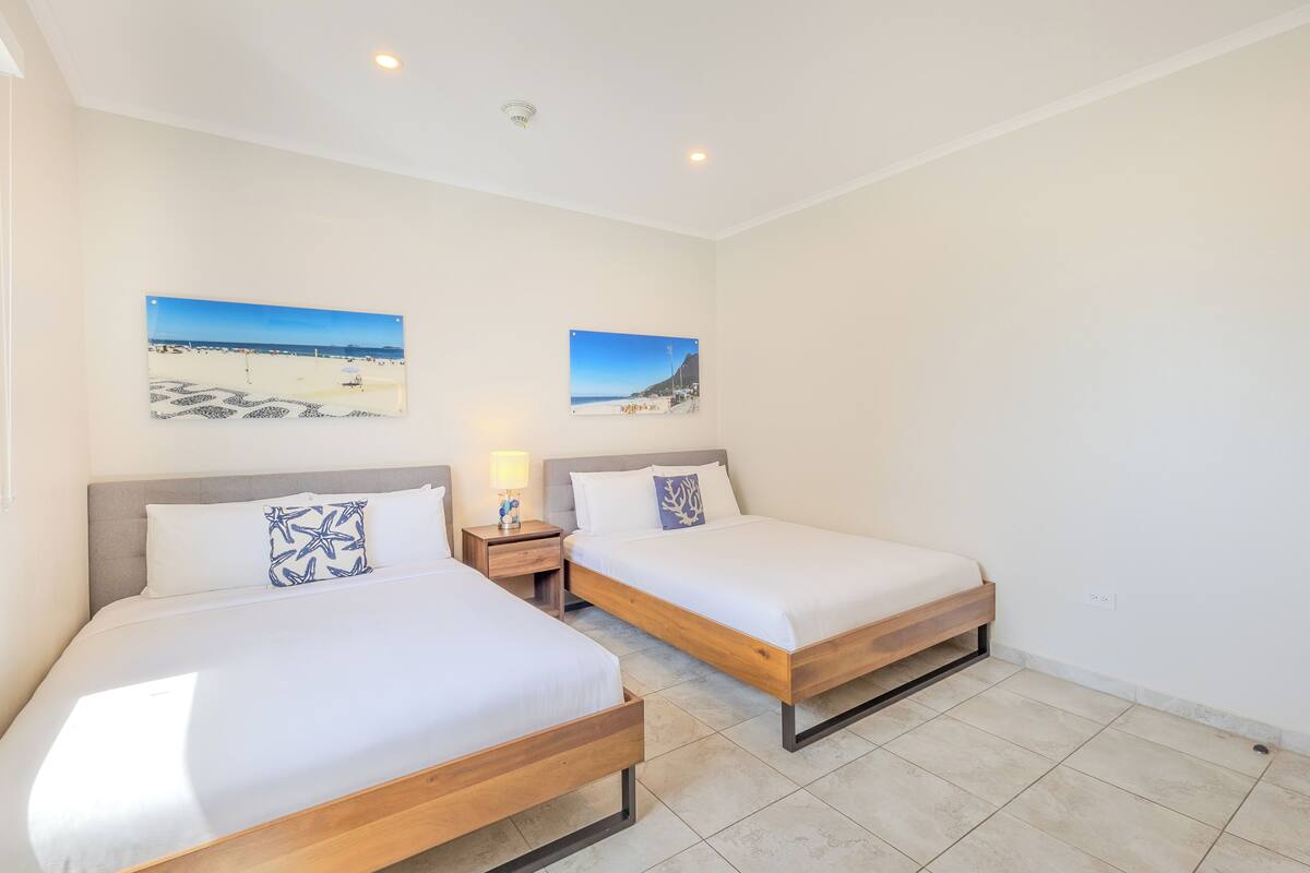 A hotel room with two beds, a nightstand with a lamp, and two beach-themed wall art pieces.