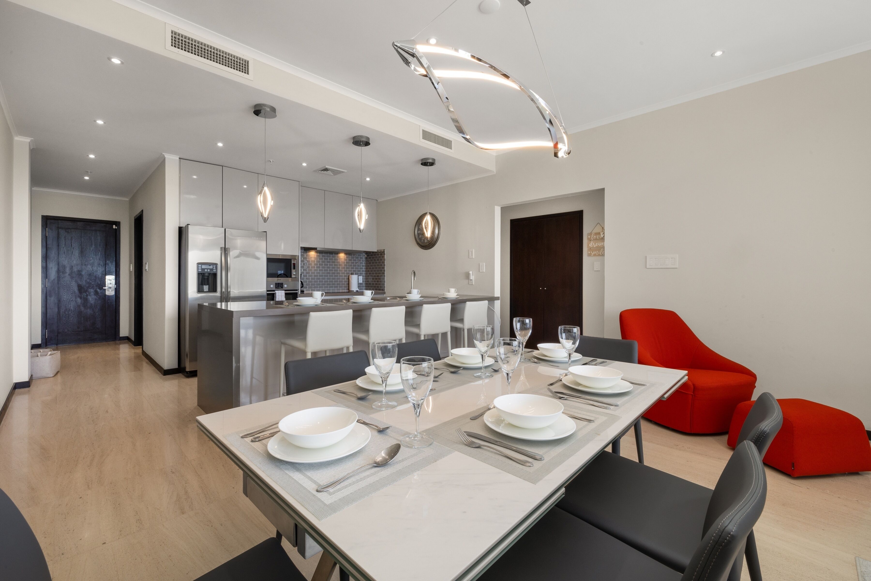 A modern kitchen with stainless steel appliances, a dining area with a long table set for six, and a red accent chair.