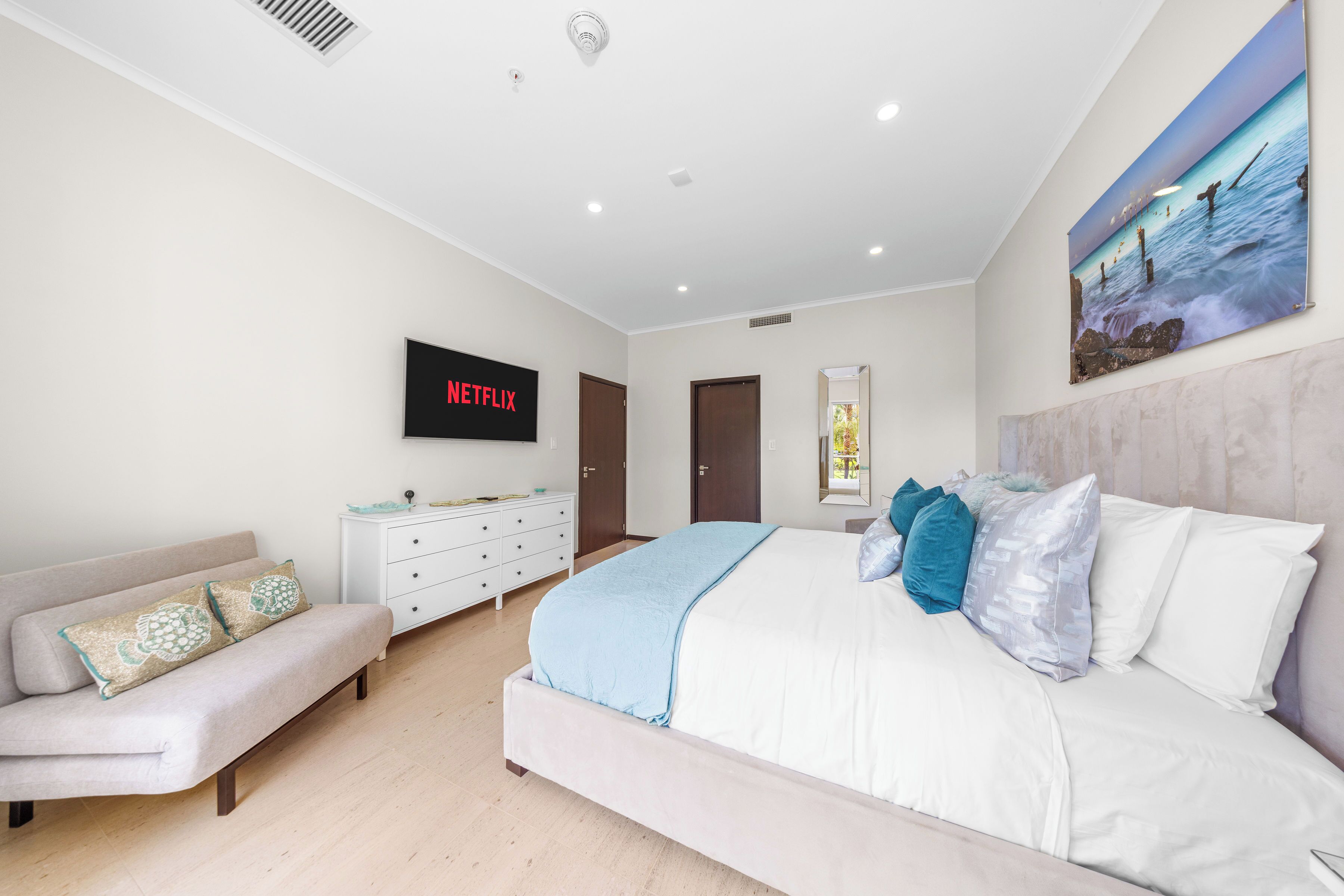 Une chambre à coucher moderne avec un lit, un canapé, une commode et un téléviseur fixé au mur qui affiche le logo de Netflix.