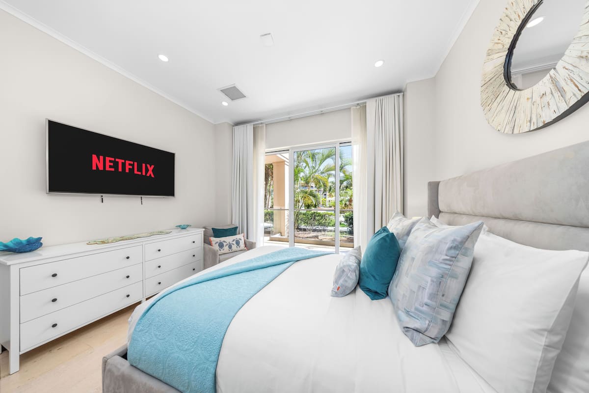 A modern bedroom with a large bed, a TV mounted on the wall, a white dresser, and a view of the outdoors through a sliding glass door.