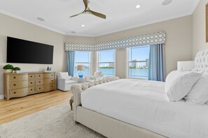 Ein Schlafzimmer mit einem großen Bett, einem Flachbildfernseher, einer Holzkommode, einem Deckenventilator und Blick aufs Wasser.