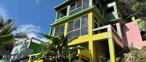 A multi-story building with a green and yellow facade, surrounded by tropical vegetation.