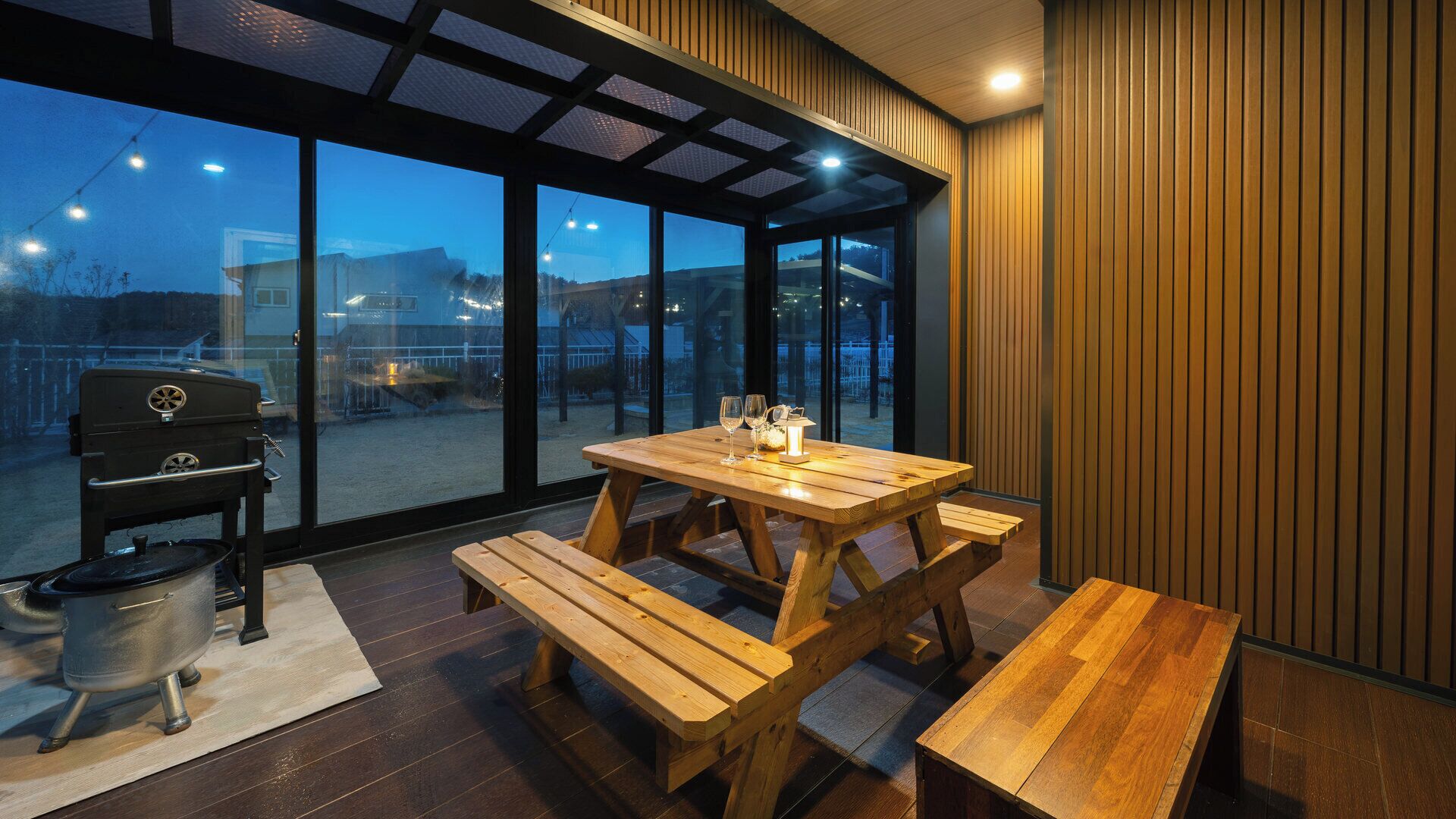 Un espacio de comedor al aire libre con una mesa y bancos de madera, una parrilla y vistas a través de amplios ventanales.
