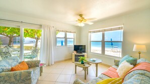Un salon avec un téléviseur, une table avec une plante et un canapé avec des coussins. Il y a des fenêtres donnant sur la plage.