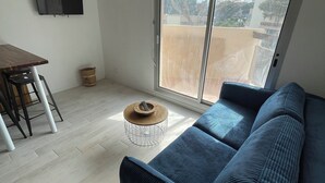 Ein modernes Wohnzimmer mit großem Fenster, einer Couch, einem kleinen runden Tisch und einem an der Wand befestigten Fernseher.