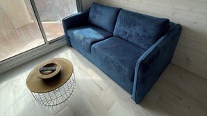 Ein modernes Wohnzimmer mit einem blauen Sofa, einem kleinen runden Couchtisch und einem großen Fenster mit Blick nach draußen.