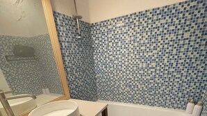Ein Badezimmer mit weißem Waschbecken, einem Spiegel und einer Badewanne. Die Wände sind mit blau-weißen Mosaikfliesen ausgekleidet.