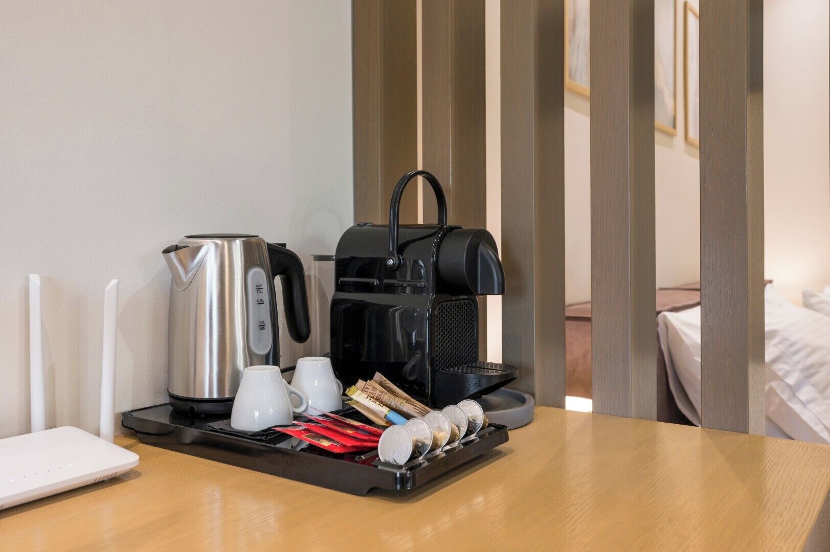 Une chambre d’hôtel avec une table en bois, une machine à café, deux tasses blanches, un plateau avec des capsules de café et un ordinateur portable.