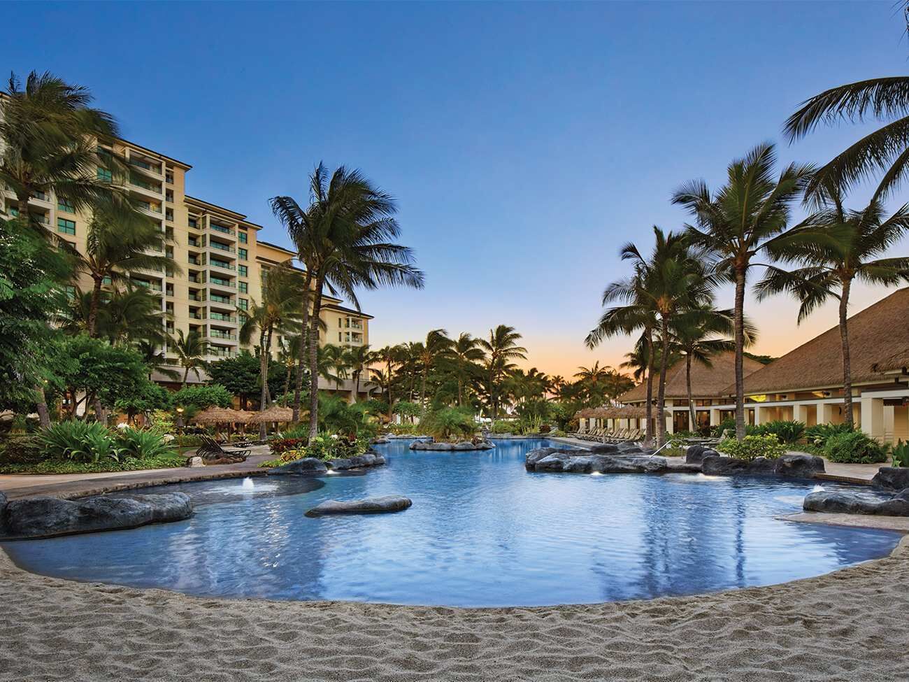 A resort pool with palm trees, a building with balconies, and a clear sky.