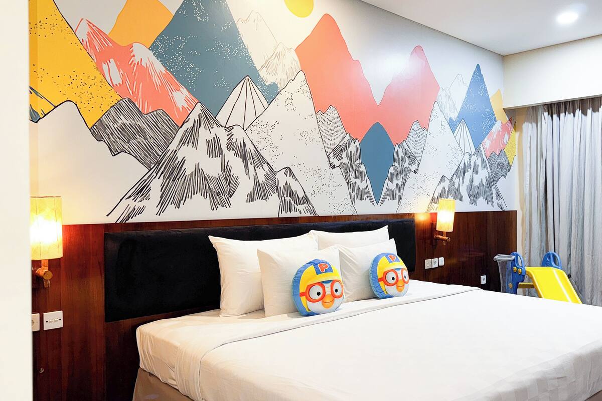 Ein Hotelzimmer mit einem großen Bett, zwei mit Zeichentrickfiguren versehenen Kissen, einem Holz-Kopfteil, einer Wandmalerei von Bergen und einem Fenster mit Vorhängen.