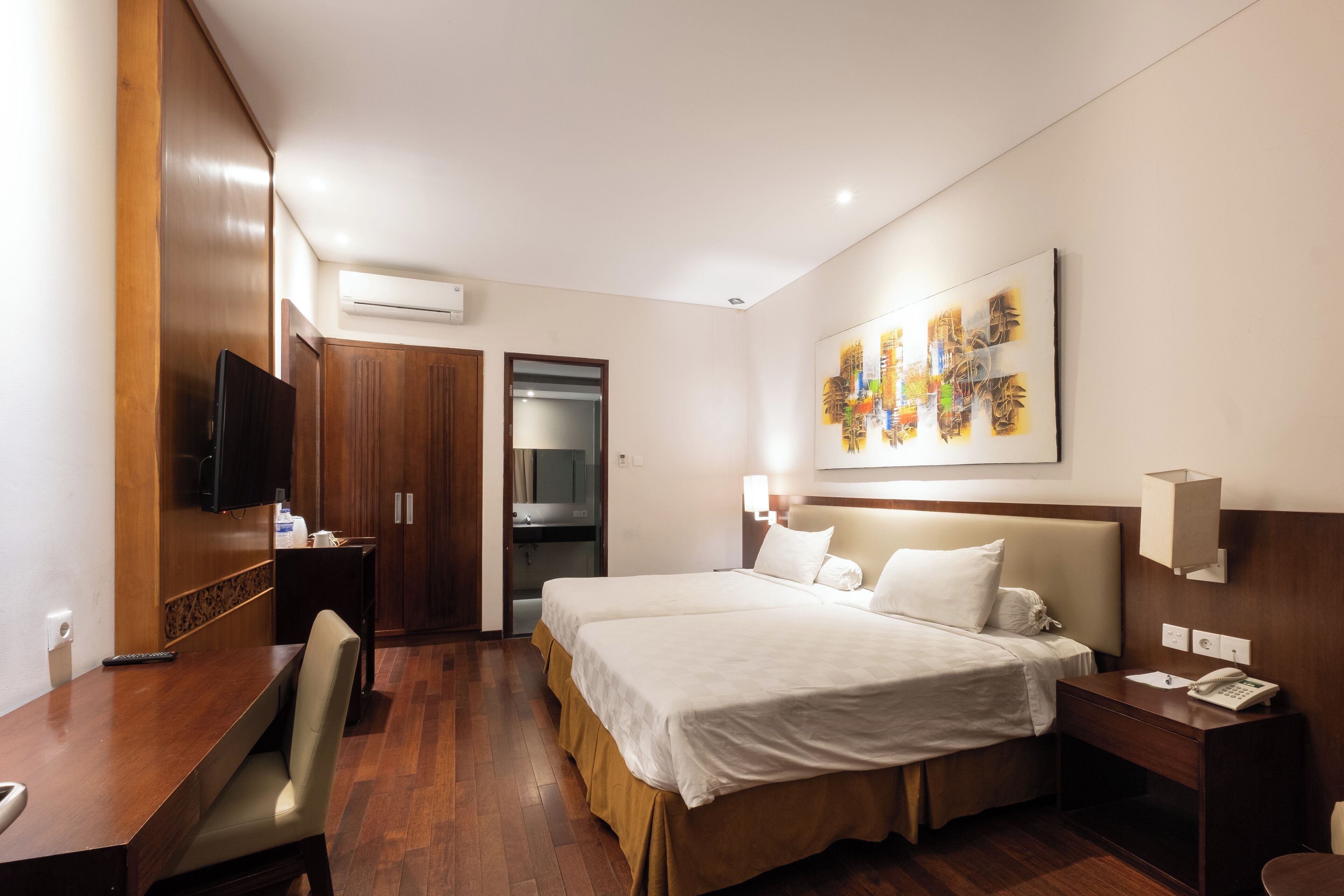 A hotel room with a bed, bedside table, desk, chair, TV, artwork, and a wardrobe.