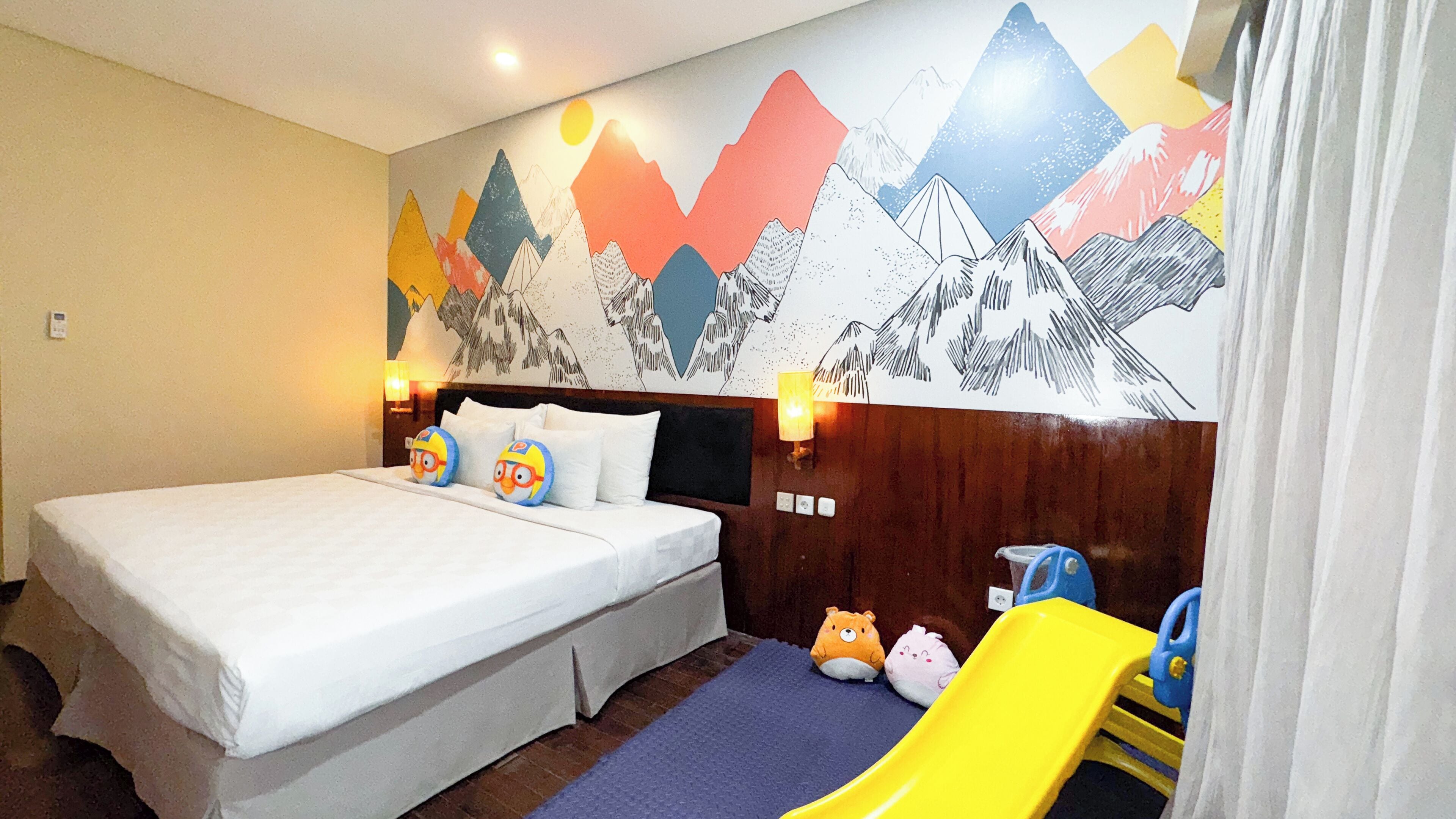 A hotel room with a bed, pillows, a nightstand, a lamp, a wall mural, a window, and a toy slide.