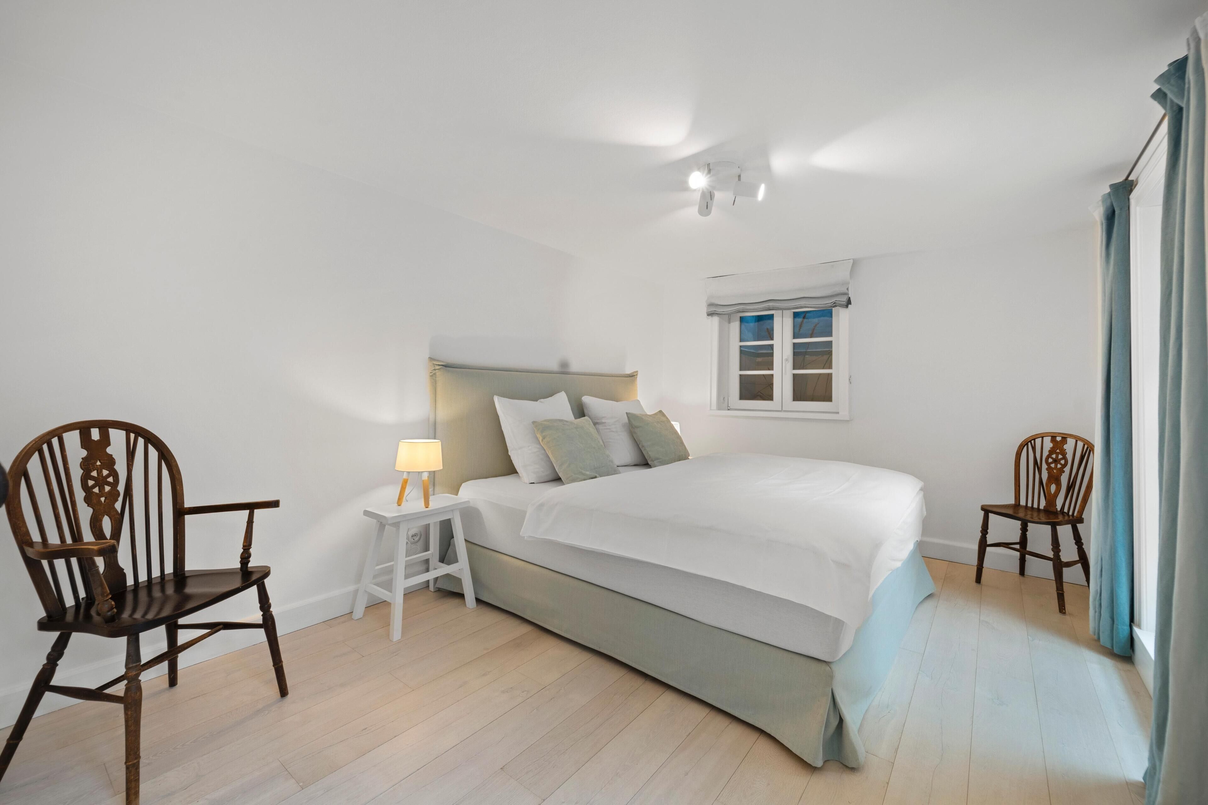 Quarto com cama, duas cadeiras de madeira, mesa de cabeceira com abajur e janela com cortinas.