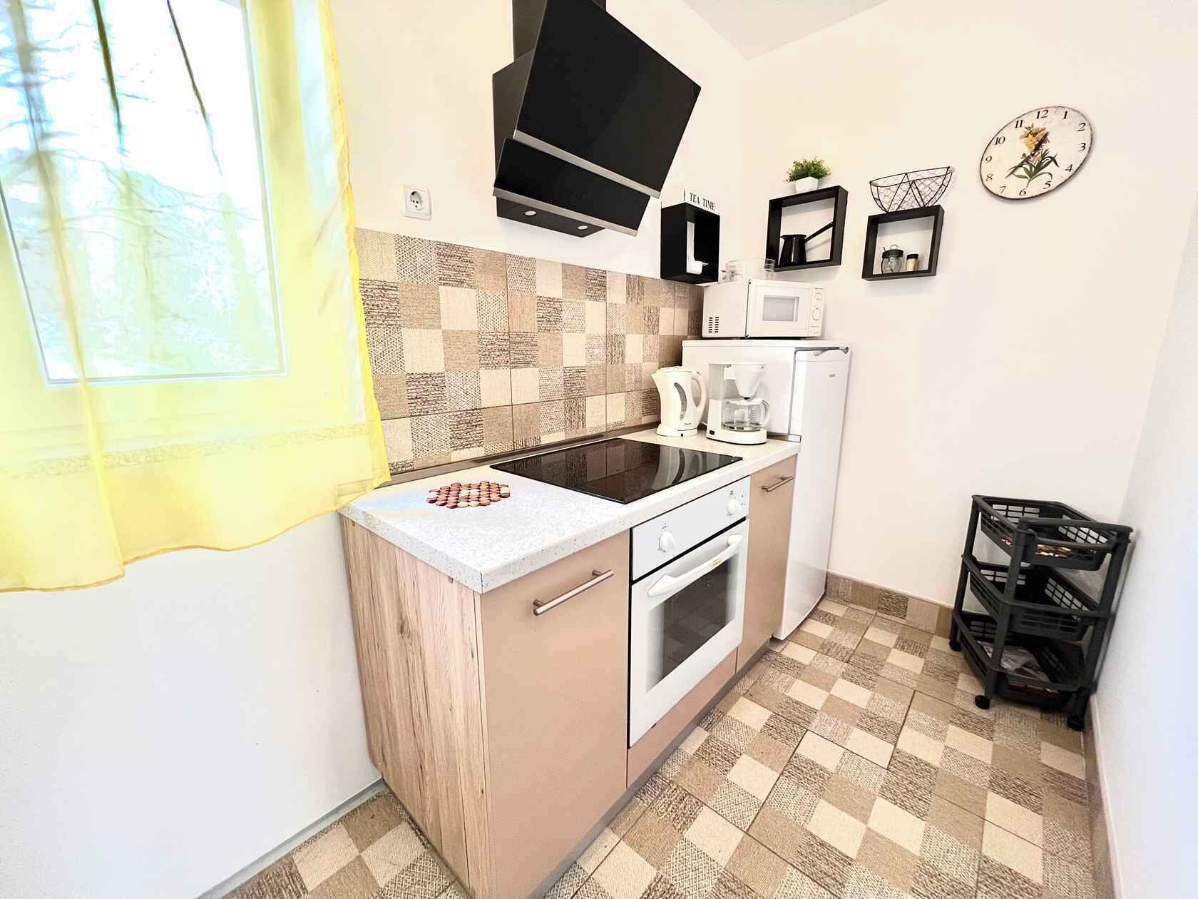 A compact kitchen with a microwave, coffee maker, and a small refrigerator. There is a window with a yellow curtain, a black TV mounted above the stove, and a clock on the wall.