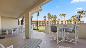 A covered outdoor patio with white furniture, including tables and chairs, and a view of a building with palm trees in the background. - Ocean Villas 19 -3 bed, 2 bath Ground floor unit just steps to the zero entry (St. Augustine Beach)