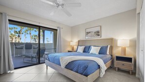 A bedroom with a bed, a ceiling fan, a nightstand, a lamp, and a view of palm trees through a sliding glass door. - Ocean Villas 19 -3 bed, 2 bath Ground floor unit just steps to the zero entry (St. Augustine Beach)