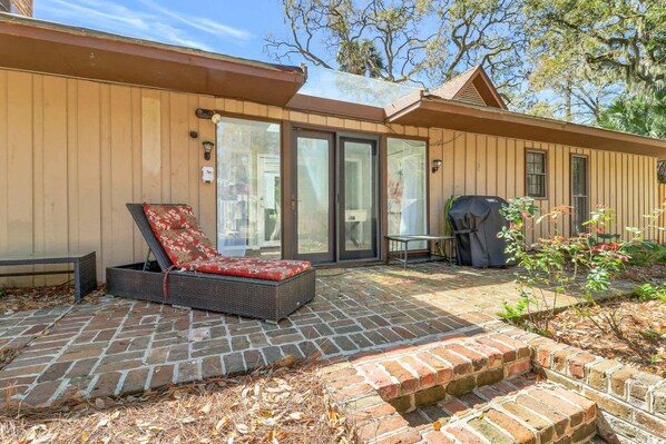 A patio area with a wicker lounge chair, a barbecue grill, and a brick pathway leading to a building with large glass doors.