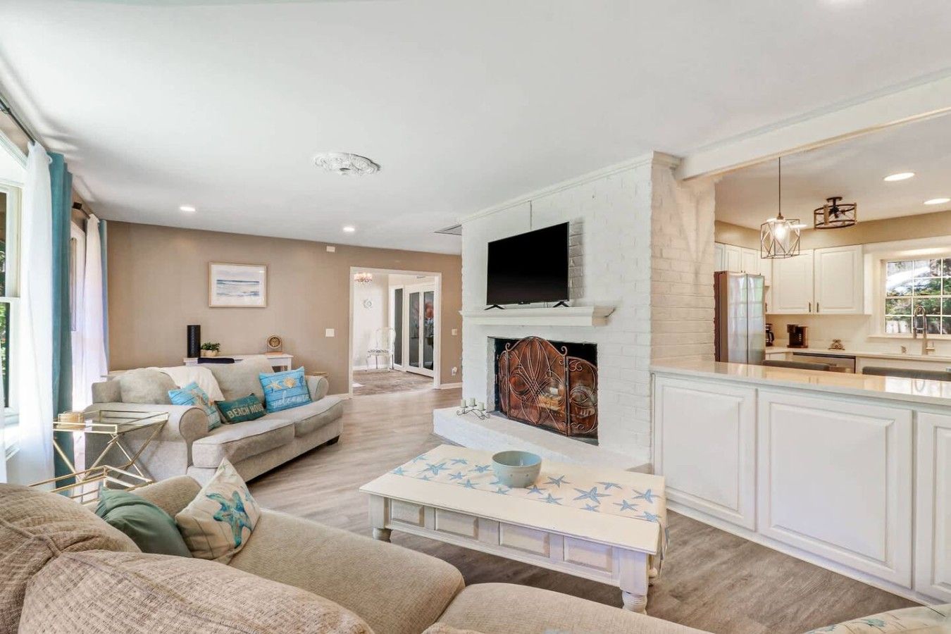A living room with a fireplace, a television, and a kitchen area with white cabinets and a window.