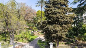Ein von einer Mauer umgebener Garten mit verschiedenen Bäumen und Sträuchern, einem gepflasterten Weg und einer Bank.