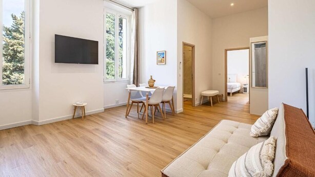 Ein geräumiger Wohnbereich mit einer Couch, einem Esstisch mit Stühlen, einem an der Wand montierten Fernseher und einem großen Fenster für natürliches Licht.